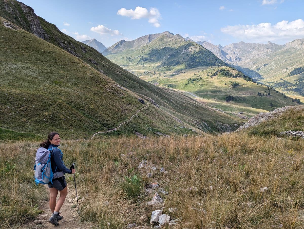 Linda wandert mit kurzer Hose und großem Wanderrucksack durch ein breites, alpines Tal, welches zu beiden Seiten von hohen Bergen flankiert wird.