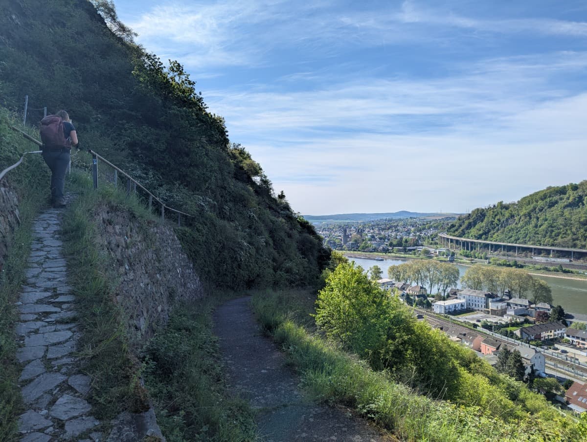 Eine Wanderin mit Rucksack steigt einen schmalen, gepflasterten Pfad am Hang entlang, der Blick reicht über eine Stadt am Rhein. Der gut gesicherte Wegverlauf mit Geländer macht deutlich, wie sich auch Anfänger sicher und mit Aussicht an das Wandern auf dem Rheinsteig herantasten können.