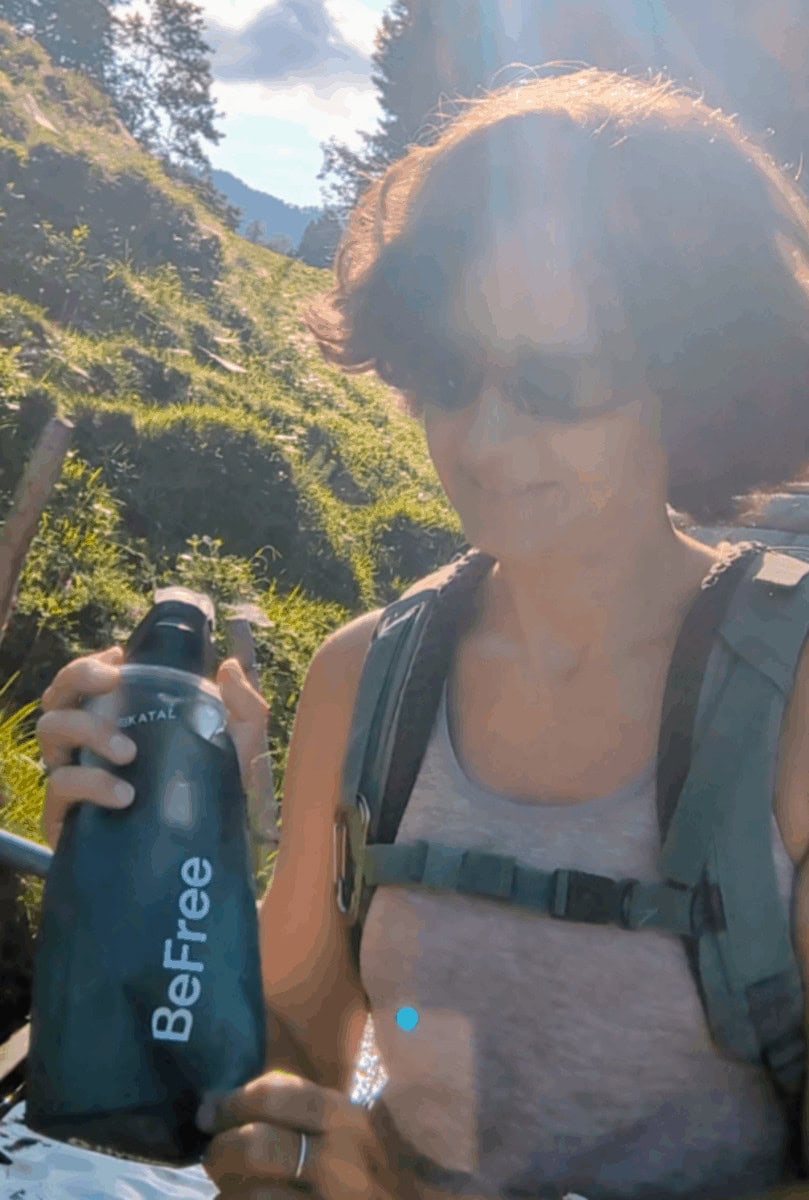 Linda präsentiert einen Trinkbeutel als Geschenk für Wanderer, während sie mit Rucksack auf einer sonnigen Almwiese steht.