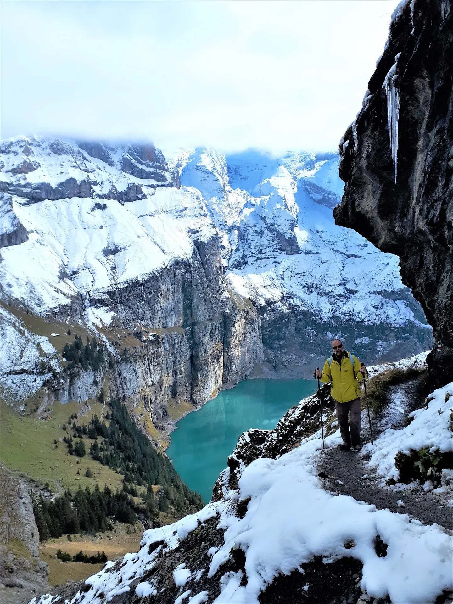 Ein Winterwanderer in gelber Jacke mit Stöcken geht auf einem schmalen, schneebedeckten Pfad an einem steilen Felshang entlang. Im Hintergrund liegt der türkisfarbene Oeschinensee eingebettet in die verschneiten Felswände der Berner Alpen, teilweise von Wolken verdeckt.