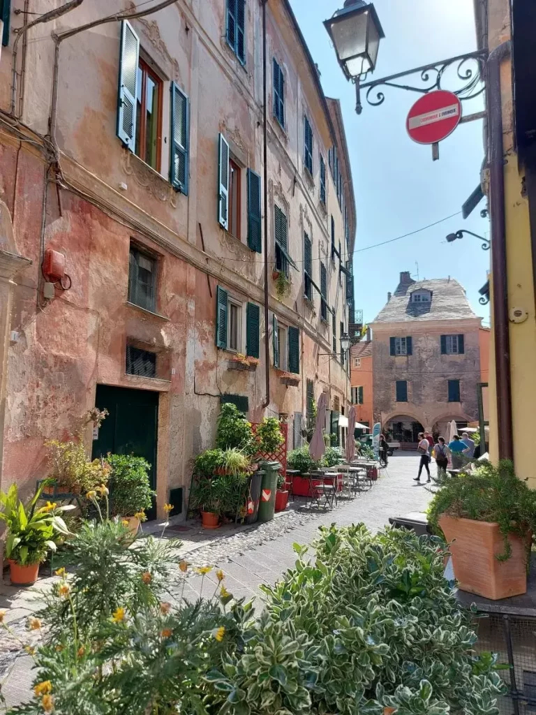 Ein charmanter Straßenzug in Finalborgo mit pastellfarbenen, leicht verwitterten Hausfassaden und grünen Fensterläden, die nostalgischen Flair versprühen. Pflanzgefäße mit üppigem Grün säumen den Gehweg, während vor einem Café Stühle und Tische einladen. Im Hintergrund führt die enge Straße zu einem kleinen Platz, wo sich Menschen zwischen historischen Gebäuden bewegen, und verleiht der Szene ein lebhaftes, mediterranes Ambiente.