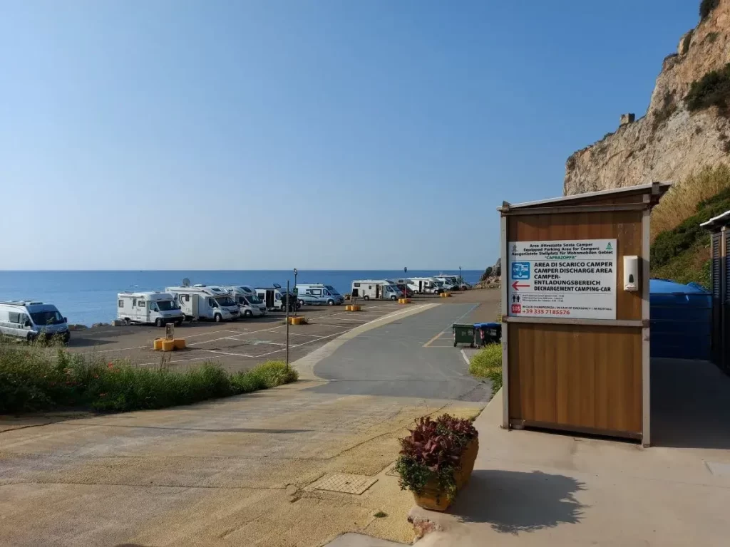 Ein Stellplatz für Wohnmobile direkt am Meer, gesäumt von geparkten Fahrzeugen, bietet Reisenden eine ruhige, panoramareiche Unterkunft. Im Vordergrund befindet sich ein Informationsschild, das Entsorgungs- und Camping-Services beschreibt, während sich im Hintergrund die felsige Küste erhebt. Die klare Sicht und das ruhige Wasser sorgen für eine entspannte Atmosphäre an diesem Küstenabschnitt.