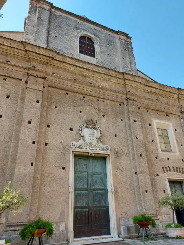Nahaufnahme einer alten Kirche in Finalborgo mit markanter, rustikaler Fassade, die viele kleine Öffnungen und Verzierungen aufweist. Der Eingang ist mit kunstvollen Reliefs geschmückt, und zwei dekorative Pflanzenrahmen flankieren die schwere Holztür. Die verwitterte Oberfläche der Kirche erzählt von einer langen Geschichte und verleiht dem Gebäude einen authentischen Charme.