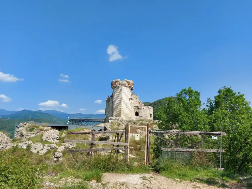 Die Burganlage oberhalb von Finalborgo zeigt alte, verfallene Mauern und eine charakteristische, runde Turmstruktur, die noch gut erhalten ist. Im Vordergrund steht ein einfacher Holzzaun und üppige Vegetation, die den Zugang zur Ruine teilweise verdecken. Der Himmel ist strahlend blau und bietet einen klaren Kontrast zu den bröckelnden weißen Steinmauern der Burg.