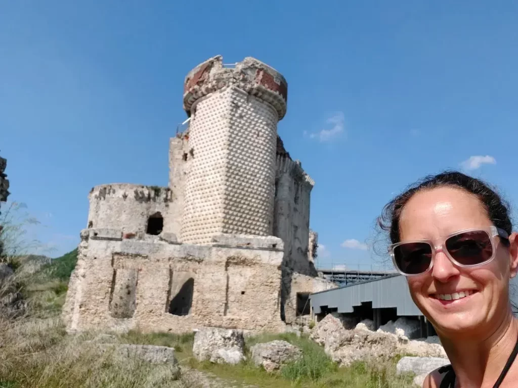 Selfie von Linda vor der Burgruine in Finalborgo, die eine mächtige, steinverzierte Struktur mit markantem Rundturm zeigt. Die Person im Vordergrund lächelt in die Kamera, während die Ruine einen faszinierenden Kontrast zur blauen Himmel bietet. Der Blick auf die Ruine vermittelt ein Gefühl von Abenteuer und Entdeckung historischer Stätten.