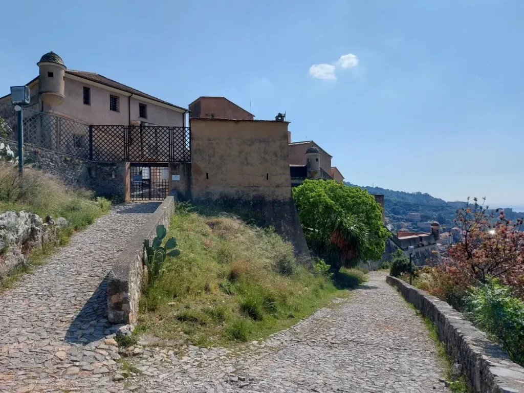 Ein steiniger Weg führt hinauf zur Festung San Giovanni in Finalborgo, flankiert von alten Gebäuden und umgeben von üppigem Grün. Das historische Mauerwerk und die Vegetation verleihen dem Weg eine romantische, verwunschene Atmosphäre. Der Weg schlängelt sich entlang der Festungsanlage und gibt Einblicke in die Geschichte der Befestigungsanlagen.