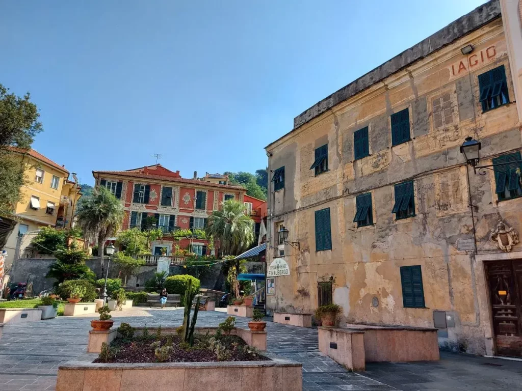Ein kleiner, ruhiger Platz in Finalborgo, umgeben von historischen Gebäuden mit abblätternder Fassade und grünen Fensterläden. Im Hintergrund hebt sich ein rot gestrichenes Gebäude mit verzierten Fensterrahmen und einem gepflegten Garten hervor. Der Platz ist mit Pflanzenkübeln und mediterranen Pflanzen dekoriert, was dem Ort eine charmante, mediterrane Atmosphäre verleiht.