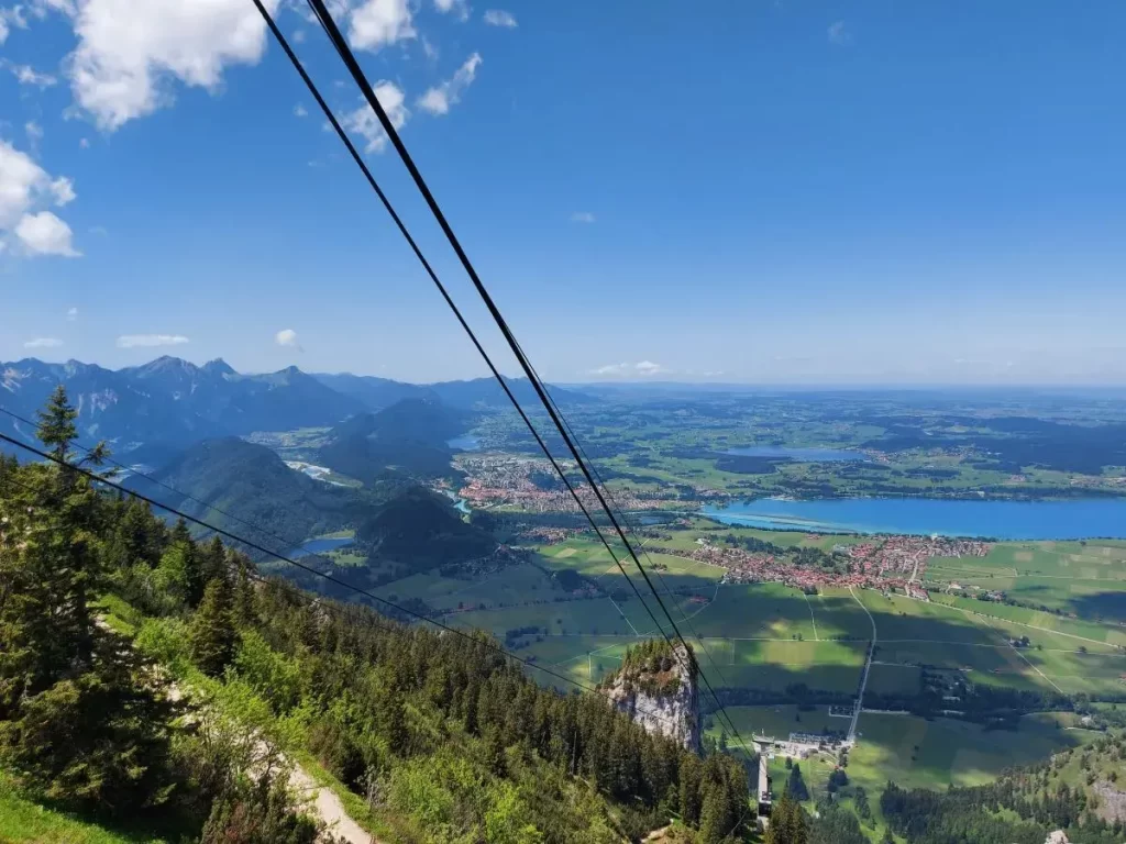 Blick von einem Wanderweg unterhalb der Bergstation der Tegelbergbahn mit Seilbahnkabeln und weitem Alpental. Dieser Abschnitt zeigt die Anbindung zwischen Bergbahn und Wanderpfad auf dem Tegelberg in Füssen.