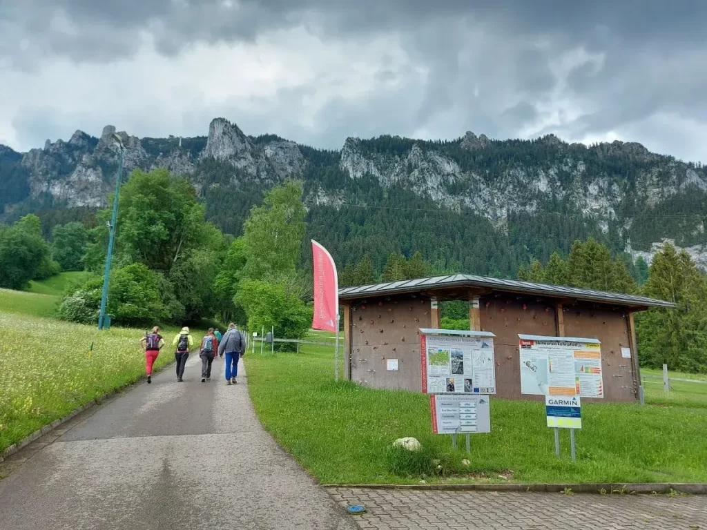 Ein Holzpavillon mit Infotafeln und Wegweisern vor steilen Felswänden des Tegelbergs. Dieses Bergsportzentrum markiert den Startpunkt für Kletter- und Wanderabenteuer in der Region rund um den Tegelberg in Füssen.