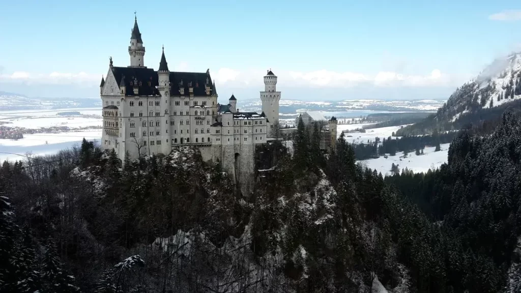 Schloss Neuschwanstein steht auf einem Felsen mit verschneiten Gipfeln im Hintergrund. Diese ikonische Aussicht von der Marienbrücke gehört zu Highlights des Tegelbergs in Füssen.