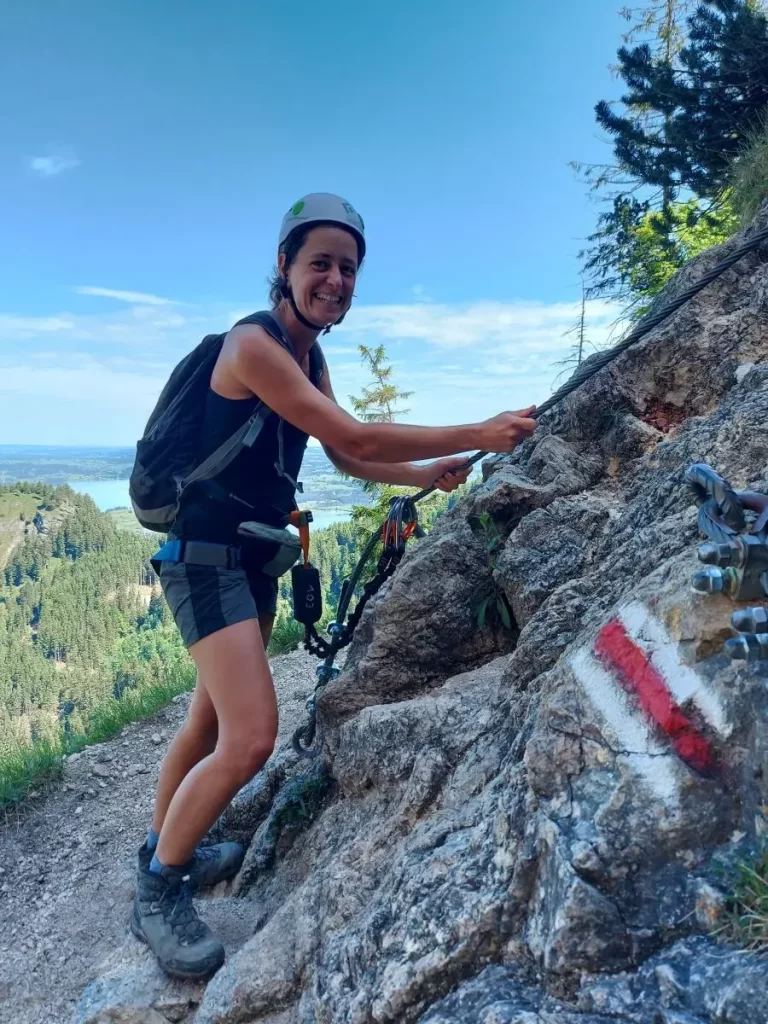 Linda trägt Helm und Klettergurt und hält sich an Sicherungsseilen am Fels. Dieses Erlebnis betont die Zugänglichkeit des Anfängersteigs Gelbe Wand am Füssener Tegelberg.