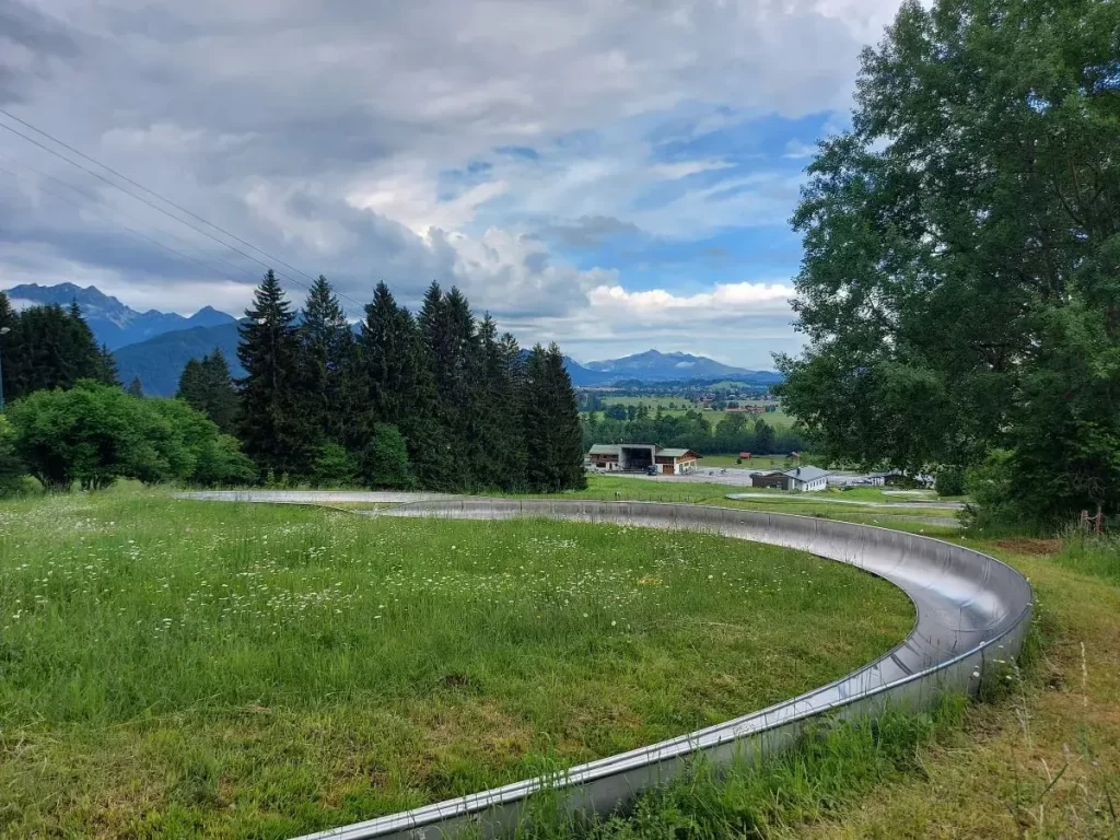 Eine kurvige Sommerrodelbahn schlängelt sich durch eine grüne Wiese vor Bergwald. Diese Attraktion bietet Familien und Wanderern eine spaßige Abfahrt nach der Tour auf den Tegelberg bei Füssen.