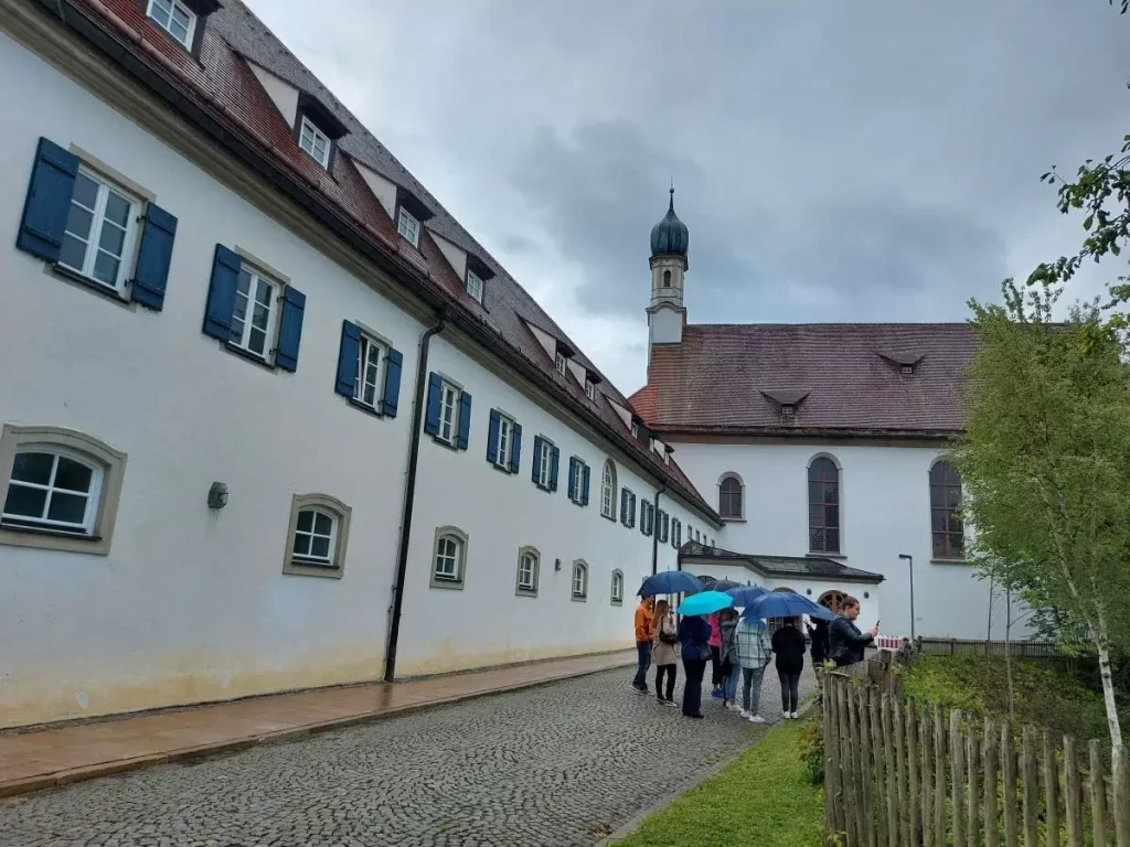 Linda und weitere Menschen mit Regenschirmen bei einer Stadtführung vor dem Franziskanerkloster St. Sebastian in Füssen