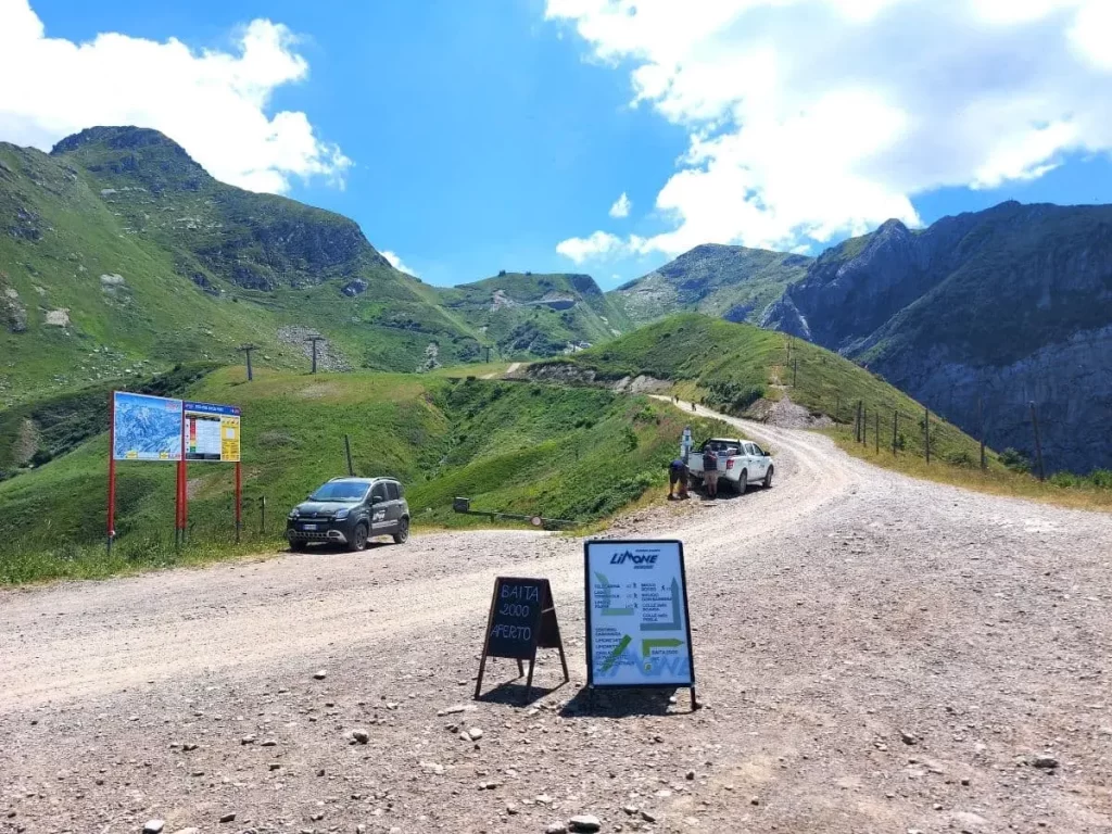 Eine staubige Bergstraße führt durch grüne Wiesen mit Parkschildern und Informationstafeln an der Seite, während ein Auto am Straßenrand parkt. Schilder wie „Baita 2000 Aperto“ deuten auf eine geöffnete Hütte hin und laden Wanderer ein. Die umliegenden Berge erheben sich majestätisch und bilden eine natürliche Kulisse für den Beginn der ligurischen Grenzkammstraße.