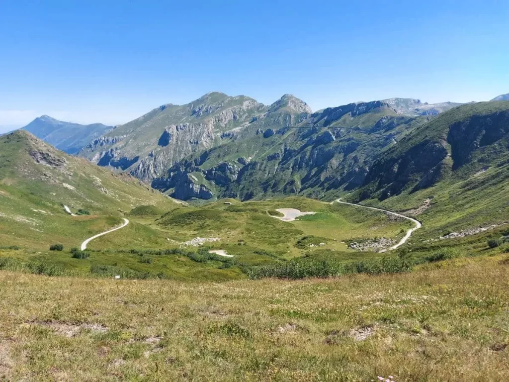 Eine kurvige Straße schlängelt sich entlang des grünen Berghangs, flankiert von steilen Felswänden und sattem Gras. Das klare Wetter erlaubt einen weiten Blick auf die angrenzenden Täler und die dahinterliegenden Berge. Diese malerische Strecke lädt zu einer Reise entlang der Grenzkammstraße ein, wo sich Natur und historische Wege vereinen.