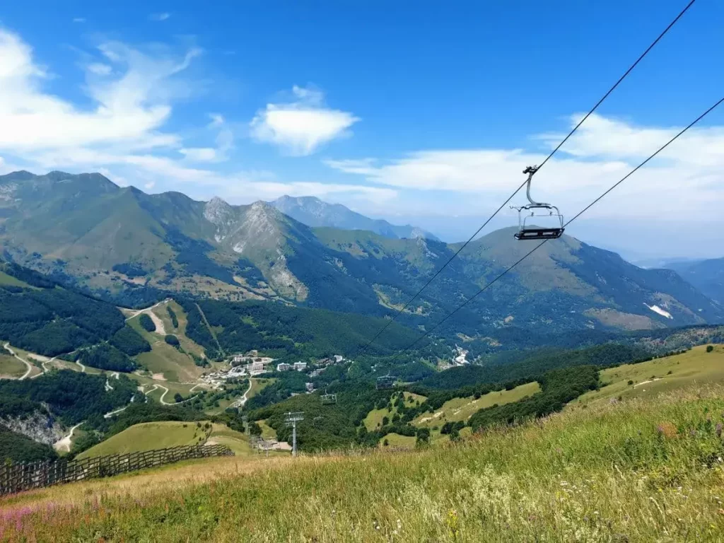 Ein Sessellift überquert eine grüne Bergwiese und führt zur Bergstation Baita 2000, die sanft in die Umgebung eingebettet ist. Der Blick schweift über das Tal und die angrenzenden Berghänge, während die klare Luft die Konturen der Berge hervorhebt. Diese Szenerie ist charakteristisch für die Erschließung alpiner Landschaften durch moderne Infrastruktur.

