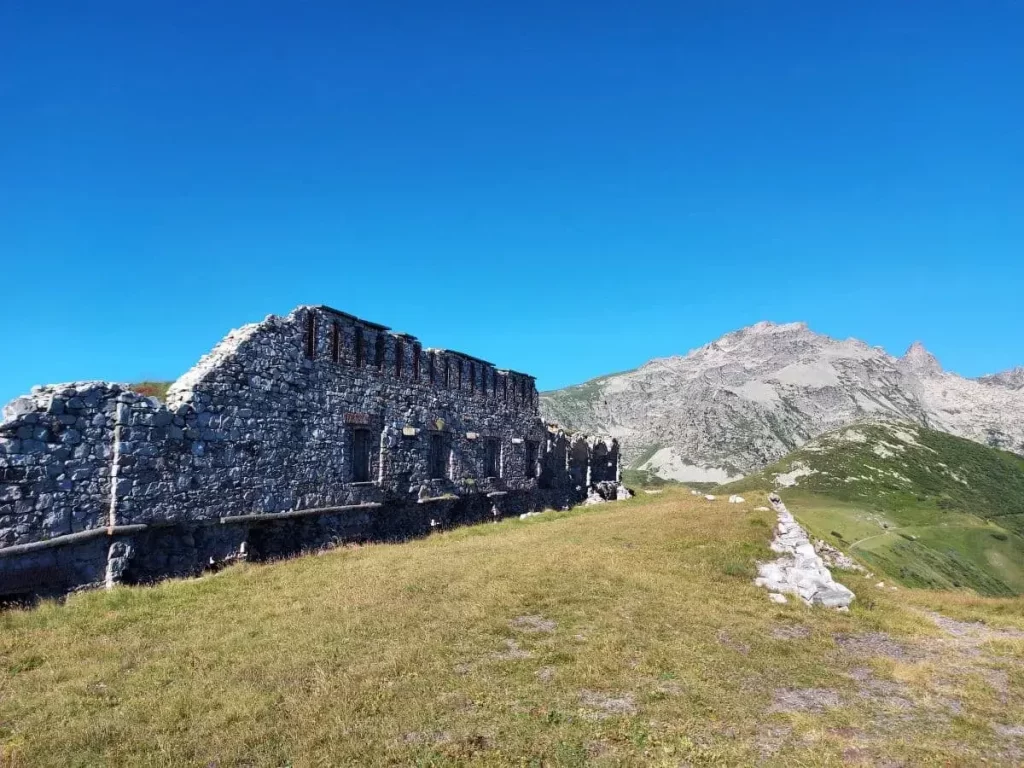 Ein verfallenes Steinfort liegt auf einer sanften, grünen Bergkuppe unter klarem, blauem Himmel. Die massiven Mauern des Fort Pernante sind von Gräsern und Wildblumen umgeben und geben den Blick auf die dahinterliegenden, schroffen Gipfel der Rocca dell'Abisso frei. Der schmale Wanderpfad führt entlang der historischen Festung und verschwindet am Horizont zwischen den alpinen Hügeln.