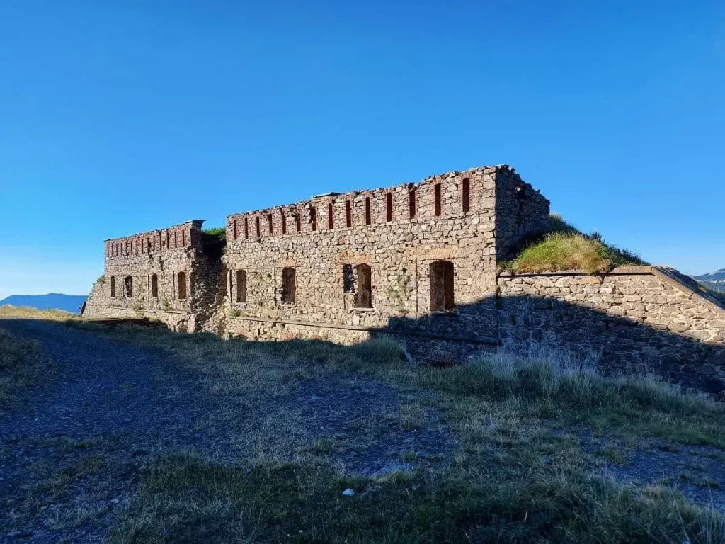 Ein massives, rechteckiges Fort aus Stein liegt verlassen auf einem grasbewachsenen Hügel am Colle di Tenda. Die verwitterten Mauern zeigen deutliche Spuren der Zeit und heben sich eindrucksvoll gegen den klaren, blauen Himmel ab. Im Vordergrund verläuft ein schmaler Weg, der um das Fort führt und die weitläufige Landschaft umrahmt.