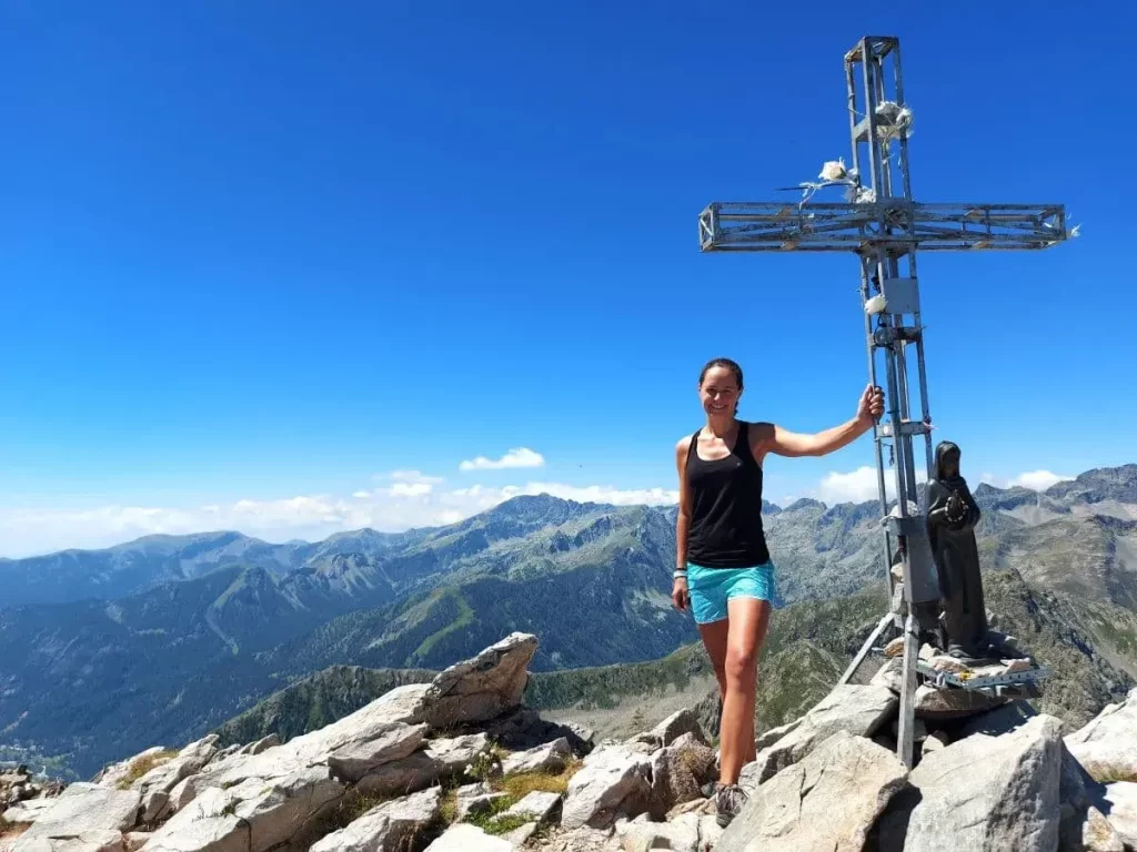 Linda in sportlicher Kleidung lehnt an einem großen, metallenen Gipfelkreuz, das hoch über den Bergen thront. Im Hintergrund erstrecken sich endlose Bergketten unter einem strahlend blauen Himmel, deren sanfte Konturen sich gegen den Horizont abzeichnen. Zu ihren Füßen ragen schroffe Felsen hervor, die die karge Schönheit des alpinen Geländes rund um die Rocca dell'Abisso betonen.