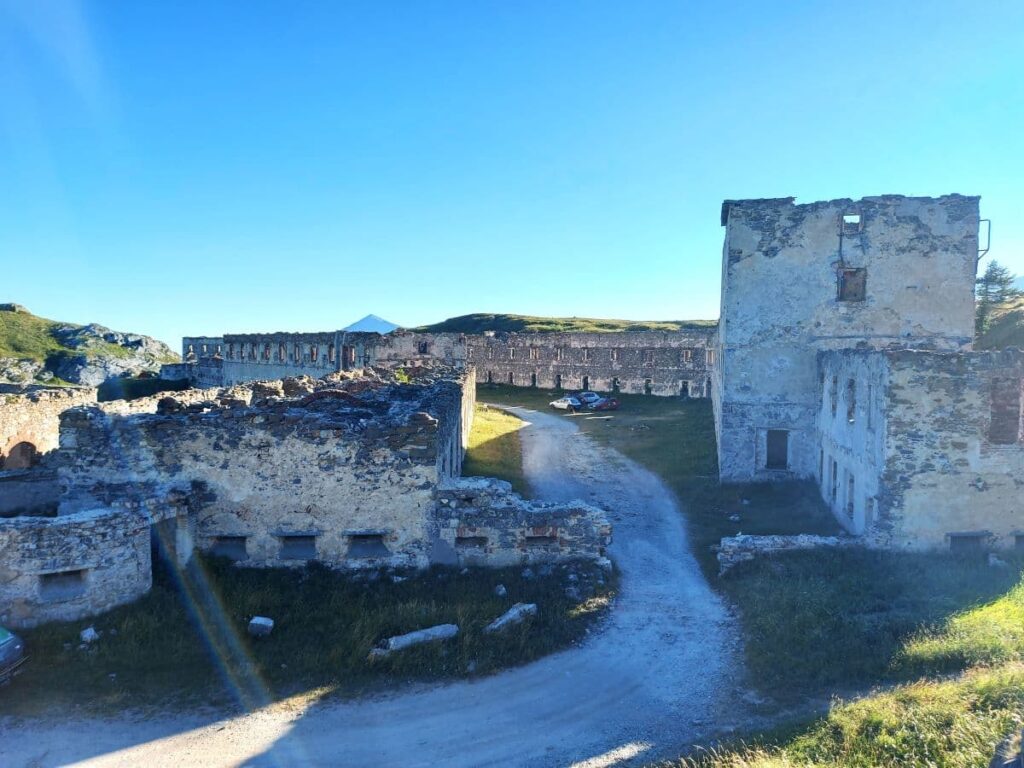 Die steinernen Mauern der Kasernen des Fort de Colle Alta stehen auf einer offenen Wiese, umgeben von sanften Berghügeln. Fensteröffnungen und verwitterte Mauerreste lassen die frühere Funktion des Bauwerks erahnen, das sich harmonisch in die alpine Landschaft fügt. Die leere Weite des Vorplatzes und der tiefblaue Himmel verstärken die majestätische, aber zugleich verlassene Atmosphäre.