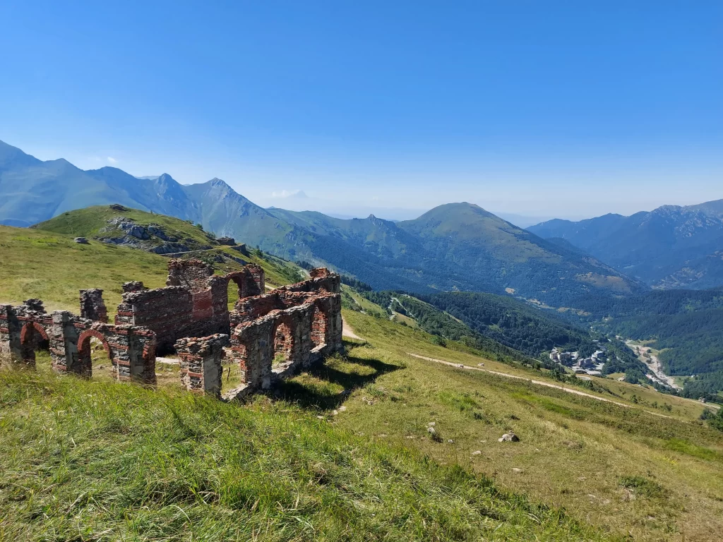 Eine freistehende Ruine mit halb zerfallenen Wänden steht auf einer Anhöhe mit freiem Blick auf das Tal. Durch die offenen Fensteröffnungen scheint das Licht und beleuchtet die Wildblumen und Gräser, die zwischen den Steinen sprießen. Die Bergkette im Hintergrund verleiht der Szenerie Tiefe und unterstreicht die Abgeschiedenheit dieser alten Stätte.