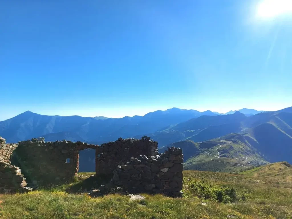 Die steinernen Überreste einer alten Festung liegen ruhig auf einer grünen Anhöhe und ragen wie stumme Zeugen der Geschichte in den Himmel. Zerfallene Mauern und Torbögen zeigen die Abnutzung der Zeit und stehen in Kontrast zur üppigen Vegetation der Umgebung. Das weite Panorama der umliegenden Berge bildet einen malerischen Hintergrund für die Ruinen.