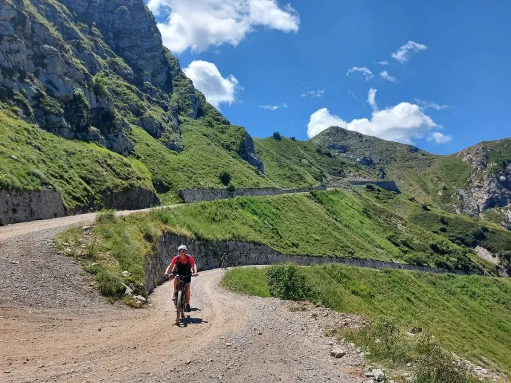 Linda auf einem Mountainbike fährt konzentriert eine Schotterstraße entlang, umgeben von saftig grünen Hängen und steilen Felsen. Der Weg schlängelt sich in sanften Kurven die Berghänge hinauf, während der klare Himmel das alpine Gelände in leuchtendem Licht erstrahlen lässt. Die Szene zeigt die Herausforderung und Freude, die die Ligurische Grenzkammstraße für Mountainbiker bietet.
