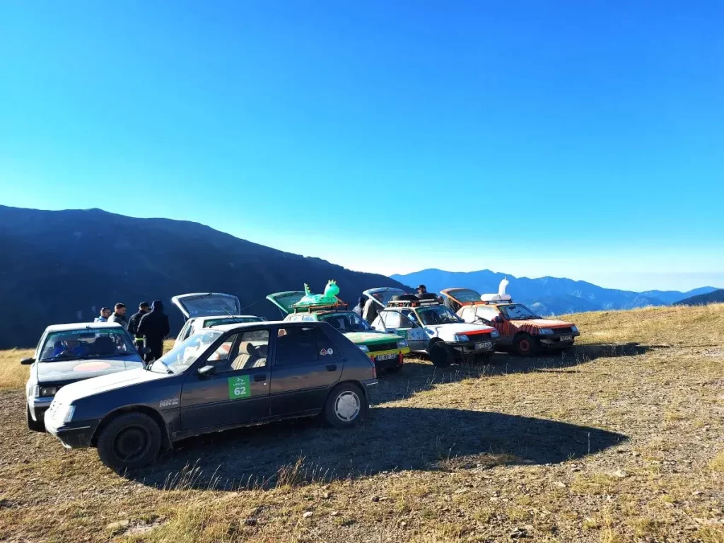 Eine Gruppe bunter Offroad-Fahrzeuge steht in einer Reihe auf einem Plateau, umgeben von weiten Bergen und klarem Himmel. Menschen versammeln sich um die Autos und genießen die entspannte Atmosphäre auf dem Colle di Tenda. Die kurvige, staubige Strecke im Hintergrund deutet auf eine abenteuerliche Fahrt durch das alpine Gelände hin.
