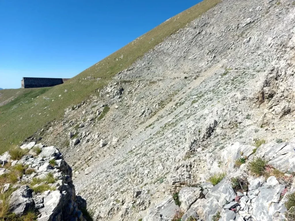 Ein schmaler, steiniger Pfad windet sich entlang eines steilen Berghangs und führt von Fort Pépin hinauf zum Grenzkamm. Die steinige Beschaffenheit des Weges und das steile Gefälle zur Seite machen die alpine Wanderung anspruchsvoll. Der Blick auf die umliegenden Gipfel und Täler verleiht der Strecke eine dramatische Note und lädt zum Erkunden ein.