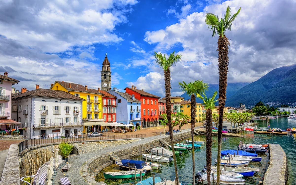 Bunte Häuserfassaden in Gelb, Rot und Blau säumen die Promenade von Ascona am Lago Maggiore, begleitet von Palmen und einem kleinen Hafen mit Booten. Im Hintergrund ragt ein Uhrturm in den dramatisch bewölkten Himmel vor der Kulisse grüner Berge.