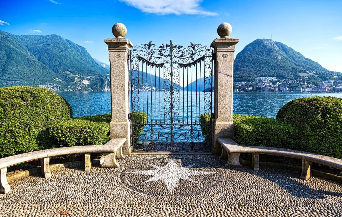 Elegantes schmiedeeisernes Tor im Parco Ciani in Lugano, flankiert von Steinbänken und gepflegten Hecken, mit Blick auf den Luganersee und die umliegenden Berge. Auf dem Boden ist ein Sternmosaik eingelassen – ein beliebter Fotospot in idyllischer Uferlage.