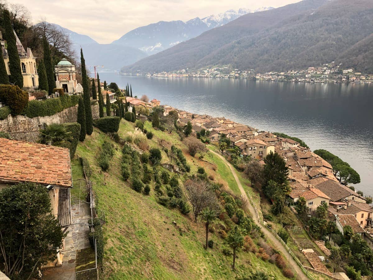 Blick auf das malerische Morcote im Tessin, das sich terrassenförmig an den Hang über dem Luganersee schmiegt. Ein absolutes Highlight im Tessin. Typische Tessiner Häuser mit roten Dächern, Zypressen und mediterraner Vegetation prägen die Szenerie, während die Alpen im Hintergrund aufragen.