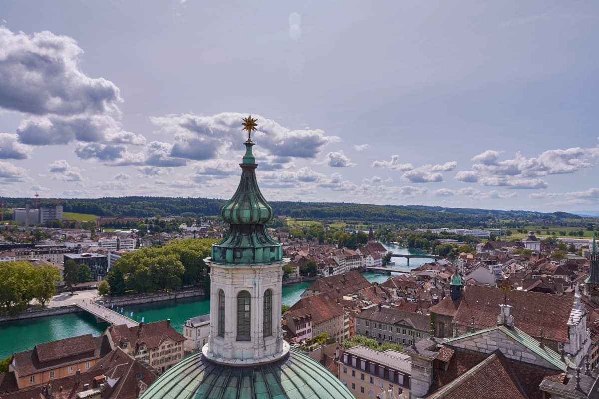 Blick von der Kathedrale St. Ursen in Solothurn über die Altstadt mit ihren roten Ziegeldächern, dem grünlich schimmernden Fluss Aare und den Brücken im Hintergrund. Die goldene Sternspitze der Kuppel thront im Vordergrund unter einem Himmel mit malerischen Wolken.
