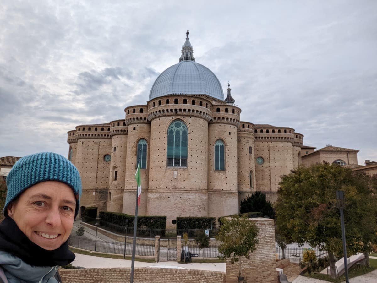 Linda lächelt in die Kamera und trägt eine türkisfarbene Strickmütze und eine Winterjacke. Im Hintergrund steht die imposante Basilika della Santa Casa in Loreto, in den Marken, mit ihrer großen silbernen Kuppel, hohen gotischen Fenstern und runden Türmen aus Ziegelstein unter einem bewölkten Himmel.