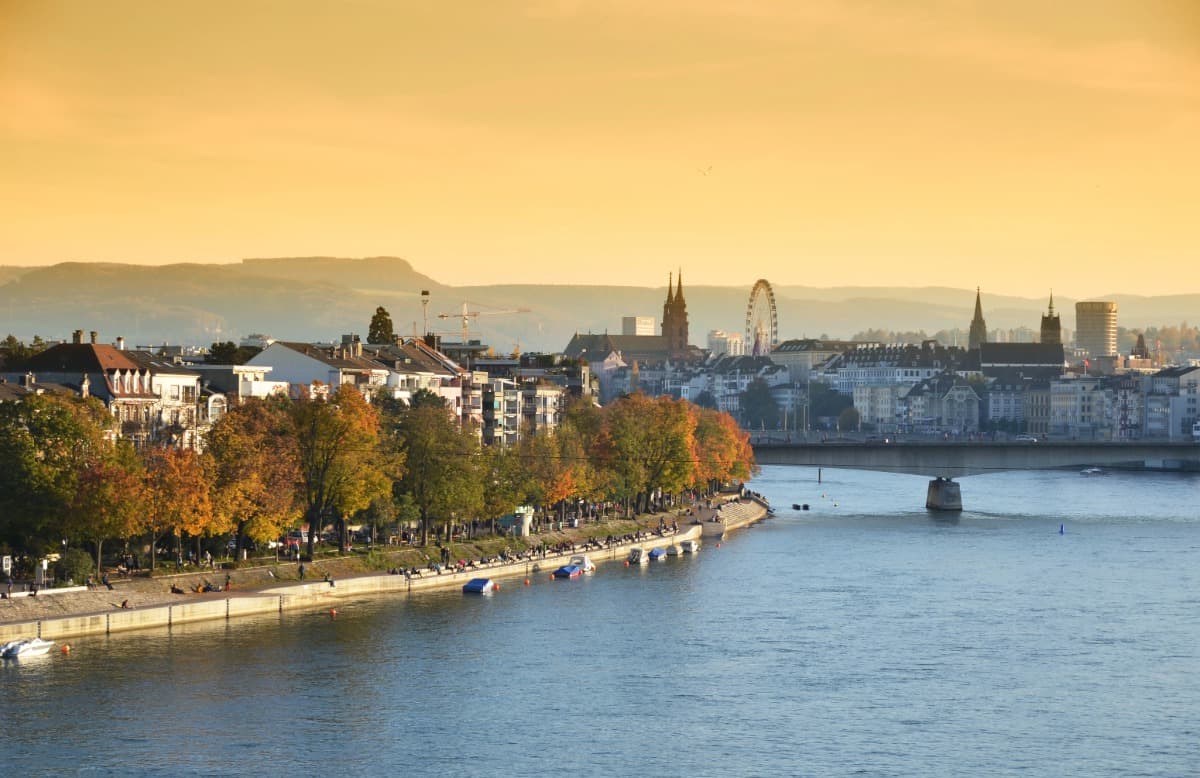 Herbstliches Panorama von Basel, Schweiz, im goldenen Abendlicht mit Blick auf den Rhein, gesäumt von bunten Bäumen und Spaziergängern am Ufer. Im Hintergrund ragen das Basler Münster, ein Riesenrad und moderne Gebäude über die Altstadtsilhouette, während eine Brücke den Rhein überspannt.