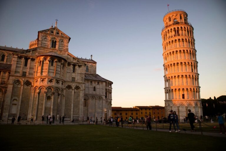 Der Schiefe Turm von Pisa und der angrenzende Dom leuchten im warmen Licht der untergehenden Sonne, während Besucher über die Rasenflächen des Campo dei Miracoli schlendern. Die berühmte Sehenswürdigkeit in der Toskana zieht zahlreiche Touristen an.
