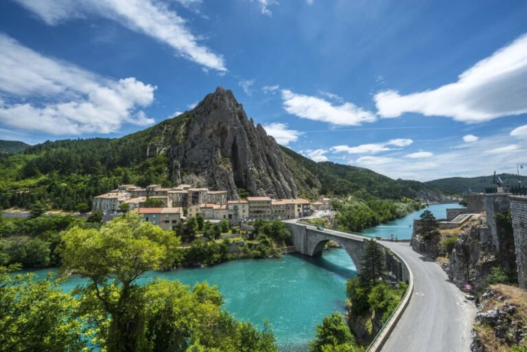 Malersicher Blick auf Sisteron an der Route Napoleon in Südfrankreich mit einer markanten Felsformation hinter dem historischen Stadtkern. Eine steinerne Bogenbrücke führt über den türkisfarbenen Fluss Durance, umgeben von grüner Landschaft und einem strahlend blauen Himmel mit weißen Wolken.