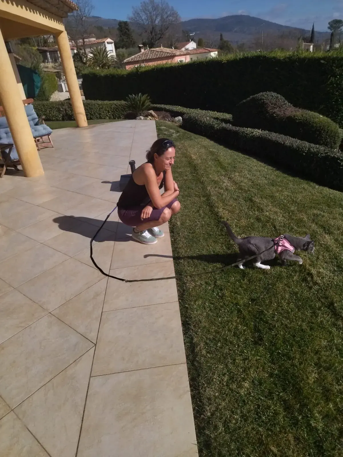 Linda hockt auf einer sonnigen Terrasse und schaut einer angeleinten Katze beim Spazierengehen über den Rasen zu. Diese Szene zeigt den Alltag als Haussitterin mit Tierbetreuung in einem privaten Zuhause.