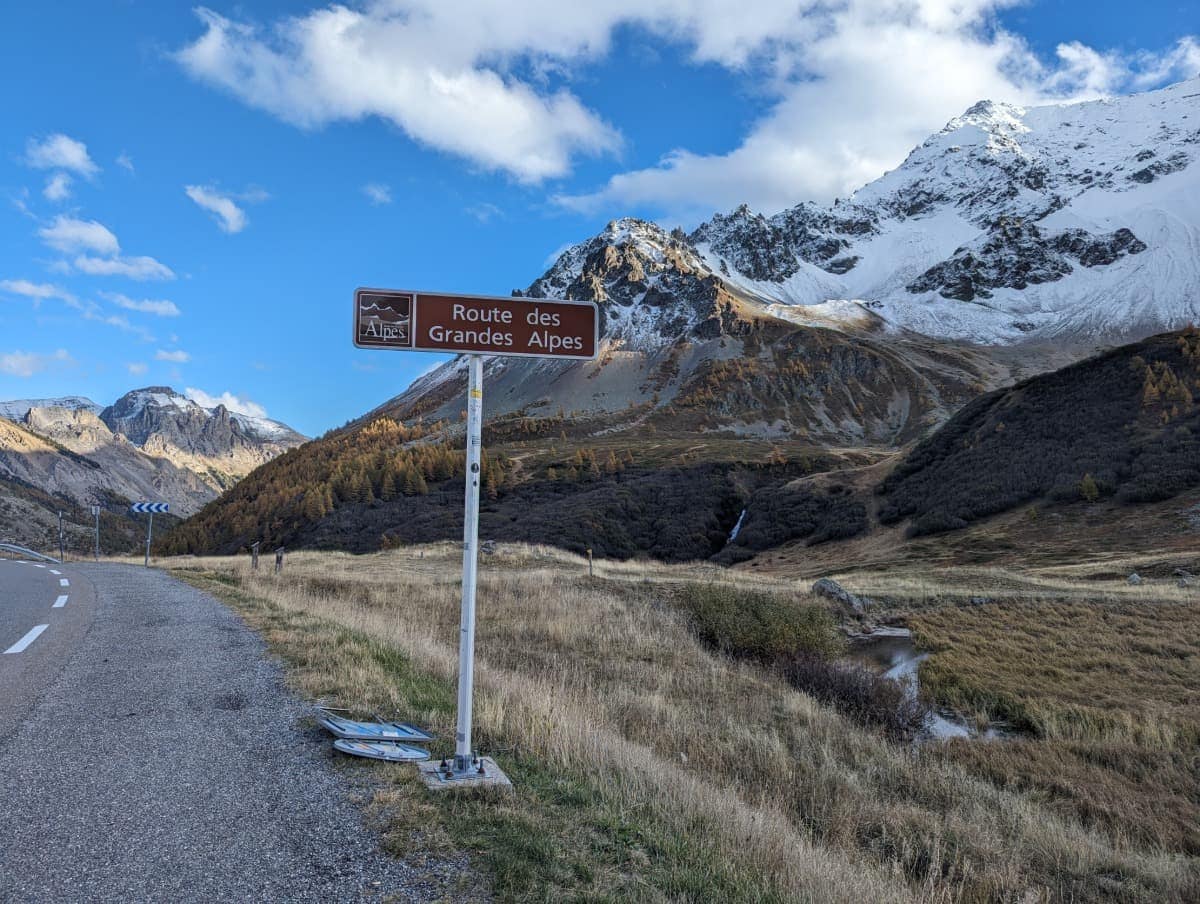 Straßenschild mit der Aufschrift „Route des Grandes Alpes“ vor einer beeindruckenden Alpenkulisse mit schneebedeckten Gipfeln, herbstlich gefärbten Bäumen und einem kleinen Bach im Tal. Die kurvige Bergstraße schlängelt sich links aus dem Bild heraus unter einem teils bewölkten, blauen Himmel.