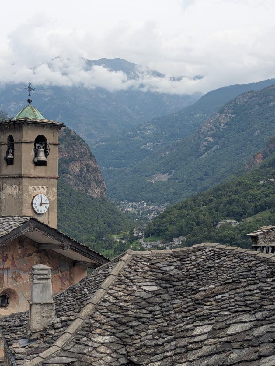 Der Kirchturm von Perloz mit Uhr und Glocken erhebt sich über den typischen Steindächern des Lystal. Im Hintergrund öffnet sich das alpine Tal mit bewaldeten Hängen und kleinen Dörfern, eingebettet in die Berglandschaft des Aostatals.