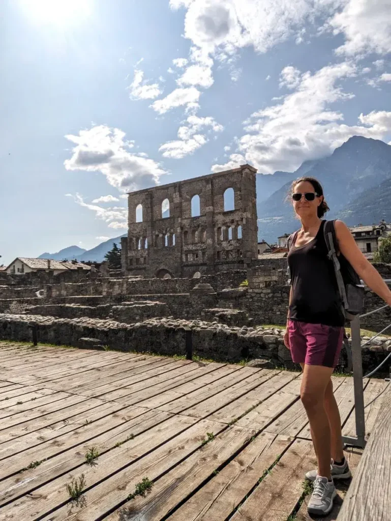 Linda steht vor den römischen Ruinen des antiken Theaters in Aosta, im Hintergrund ragen die Alpen in den Himmel. Die Aufnahme verbindet Kulturgeschichte mit dem alpinen Panorama und passt ideal zum Thema Sehenswürdigkeiten im Aostatal.