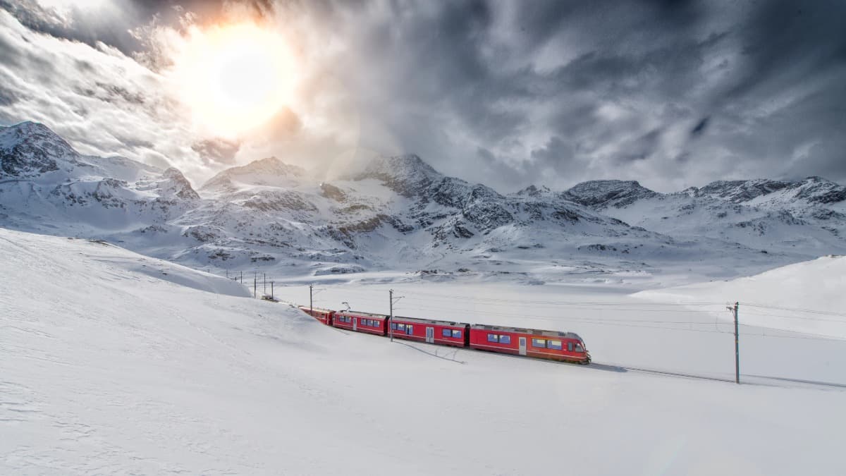 Der Bernina Express, ein tolles Ausflugsziel in der Schweiz im Winter, fährt durch eine schneebedeckte Winterlandschaft, umgeben von majestätischen Berggipfeln. Die roten Waggons heben sich kontrastreich vom weißen Schnee ab, während die Sonne dramatisch durch dichte Wolken am Himmel scheint. Ein beeindruckendes Panorama, das die alpine Schönheit perfekt einfängt.