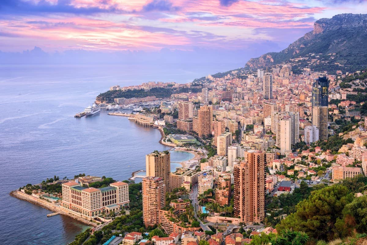 Panoramablick auf Monte Carlo in Monaco bei Sonnenaufgang, mit hohen Wohn- und Hotelgebäuden, dem Yachthafen und der Küstenlinie entlang des Mittelmeers. Der Himmel ist in sanften Rosa- und Lilatönen gefärbt, während das Meer ruhig in der Morgendämmerung liegt. Ein Muss für Ausflüge von Nizza aus