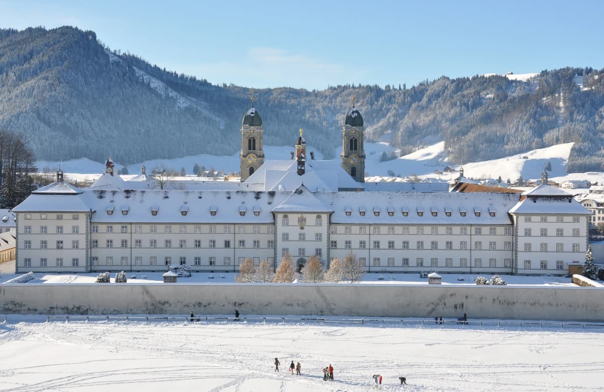 Barocke Klosteranlage von Einsiedeln in der Schweiz, eingeschneit und vor einer bewaldeten, winterlichen Hügellandschaft gelegen. Im Vordergrund spielen Menschen im Schnee auf dem Klosterplatz unter strahlend blauem Himmel.