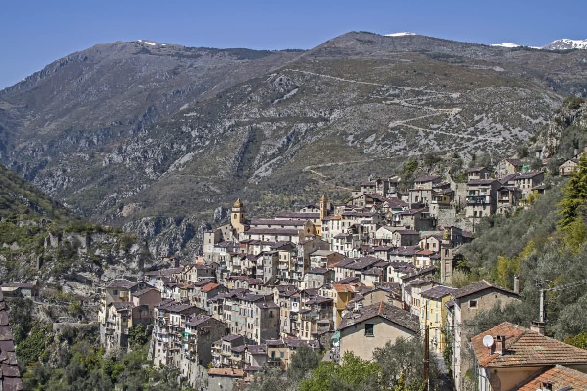 Das Bergdorf Saorge im französischen Royatal schmiegt sich terrassenartig an den steilen Hang, mit eng stehenden Steinhäusern und mehreren Kirchtürmen. Im Hintergrund ragen die felsigen Alpenhänge empor, teils mit Schneeresten bedeckt, unter einem klaren blauen Himmel. Ein tolles Ausflugsziel ab Nizza.