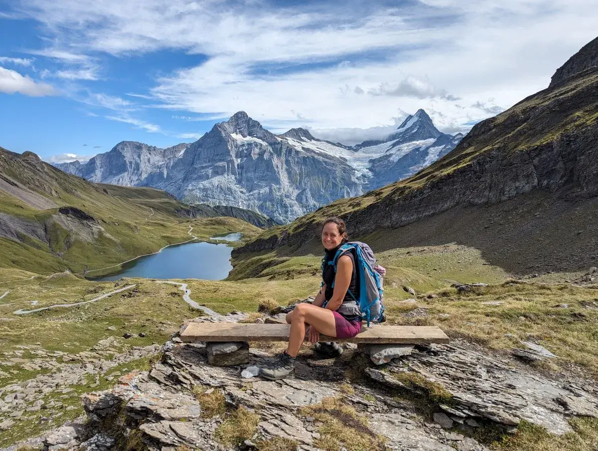 Linda sitzt auf einer Holzbank am Bachalpsee auf Etappe 15 der ViaBerna. Im Hintergrund die Gipfel des Jungfraumassiv.