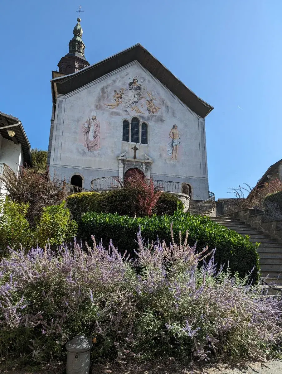Bemalte Kirche Saint-Gras d'Aosta von aussen. Davor wachsen bunte Blumen und eine breite Treppe führt zum Eingang