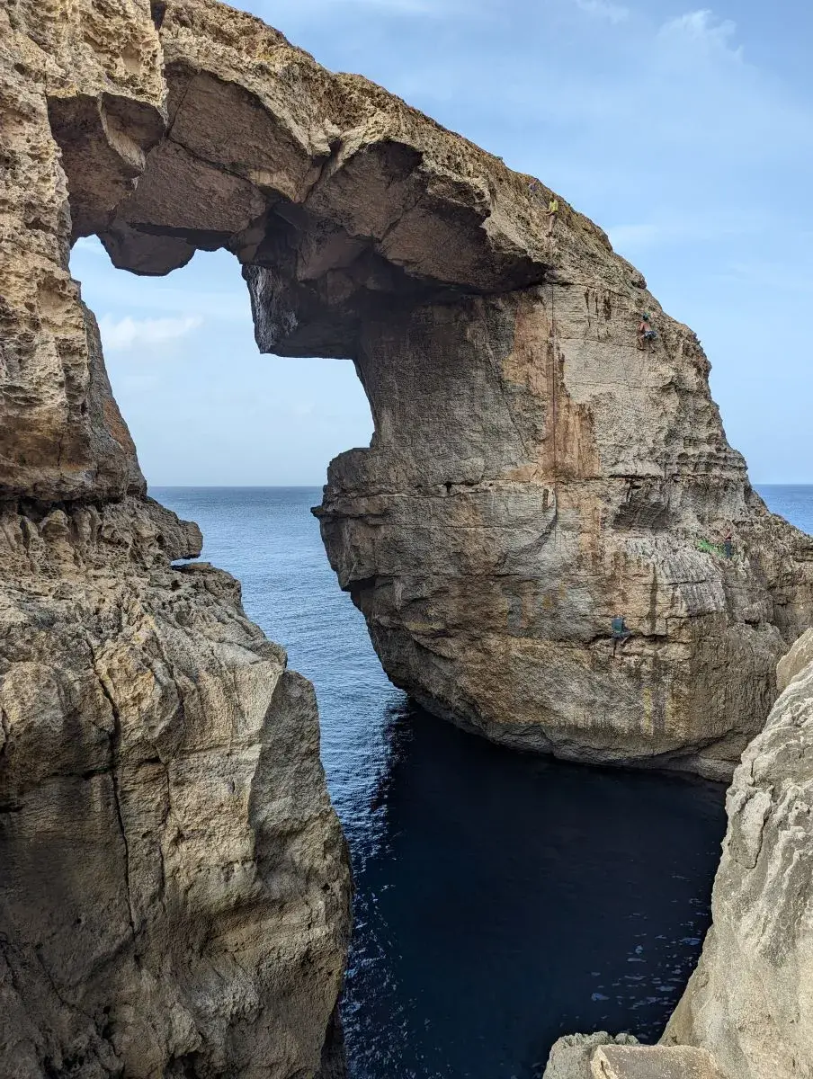 Das majestätische Wied il-Mielaħ Felstor auf Gozo. Ein natürlicher Kalksteinbogen über dem tiefblauen Meer, dessen hoch aufragende Klippen von Kletterern unter einem teilweise bewölkten Himmel erklommen werden.