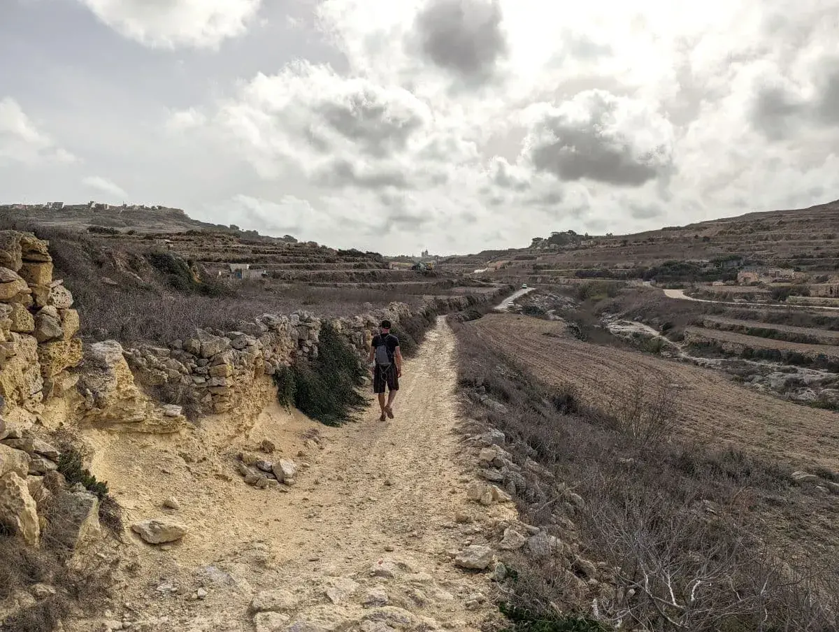 Jan wandert auf einem staubigen, von traditionellen Trockenmauern gesäumten Weg auf dem Gozo Coastal Walk. Terrassenfelder und eine ländliche Landschaft, die sich unter einem bewölkten Himmel in die Ferne erstreckt.