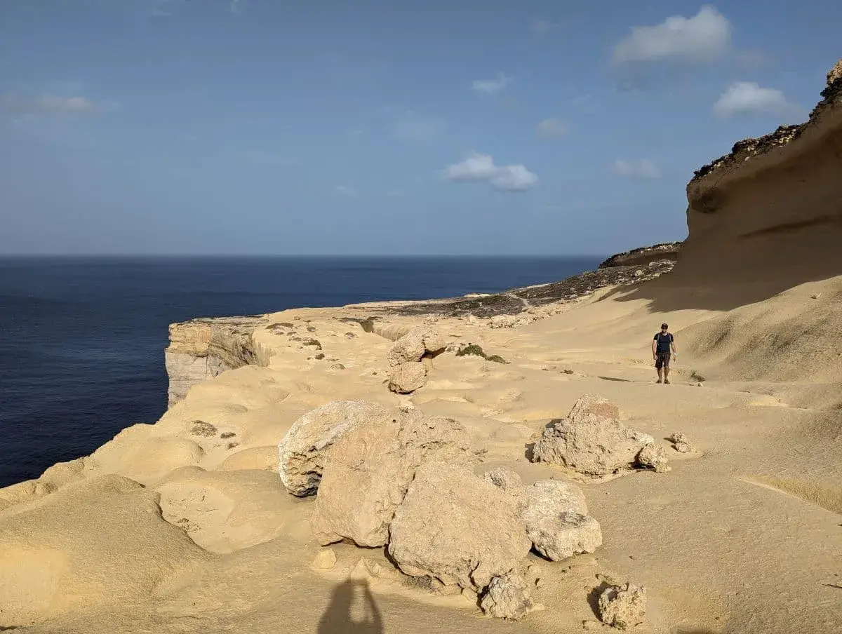 Linda durchquert das zerklüftete, sandige Terrain entlang der steilen Kalksteinklippen der Küste Gozos, während sich das weite, ruhige blaue Mittelmeer bis zum Horizont erstreckt.